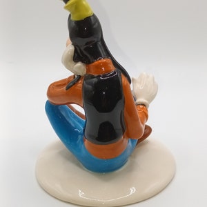 May include: A ceramic figurine of Goofy sitting with his legs crossed, wearing a blue shirt, orange trousers, and a black waistcoat. He has a yellow hat and a white glove on his right hand.
