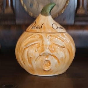 May include: A ceramic onion-shaped jar with a green stem. The jar is tan with a face carved into the front and the words "Pickled Onions" written on the side. The jar has a lid.