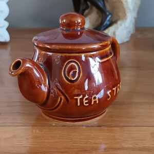 May include: A small, glossy brown ceramic teapot with a matching lid. The teapot features a rounded body, a short spout, and embossed lettering on the side that reads TEA STASH. The surface has a reflective, glazed finish with a smooth texture.