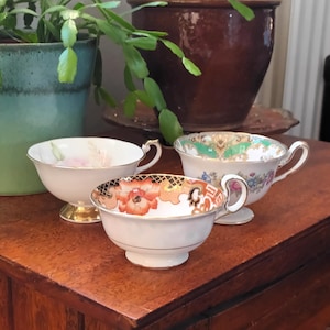 Op de afbeelding: Drie vintage theekopjes met bloemmotieven. De theekopjes zijn wit met gouden randjes en staan op een houten tafel.
