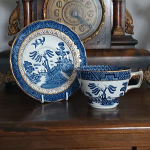 Könnte beinhalten: Blau-weißes Porzellan-Teeservice mit Landschaftsmotiv und Goldrand. Das Set wird auf einer dunklen Holzoberfläche präsentiert. Eine antike Uhr befindet sich im Hintergrund.