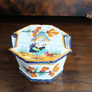 Peut inclure: Un récipient en céramique blanche avec couvercle de forme octogonale, orné de motifs floraux peints à la main en orange et vert, et d'une illustration centrale d'une personne jouant de la flûte. Le couvercle a une anse rayée bleu et blanc.