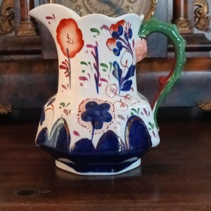 May include: A white ceramic pitcher with a scalloped rim, adorned with hand-painted floral designs in red, blue, and green. The handle is green with a red accent. The pitcher sits on a dark wooden surface.