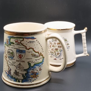 May include: Two white ceramic mugs with gold trim. One mug features a map of Devon, England, with the text "Devon" and "Dartmoor" printed on it. The other mug features a map of Somerset, England, with the text "Somerset" printed on it.