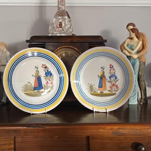 May include: Two antique ceramic plates with a white base, blue and yellow striped rims, and a central illustration of a woman in traditional attire. The plates are displayed on a wooden surface.