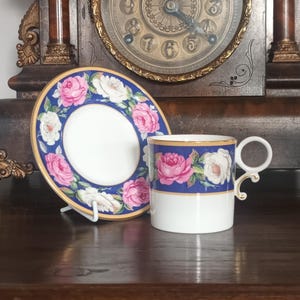 May include: A porcelain teacup and saucer set. The cup and saucer feature a blue border with pink and white rose floral designs. The cup has a white handle and a gold trim. The set is displayed on a wooden surface.