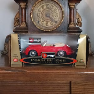 以下が含まれることがあります： ディスプレイボックスに入った赤いポルシェ356Bカブリオレ1961モデルカー。モデルは1/24スケールです。「Burago」のロゴと「Porsche 356B Cabriolet 1961」の文字が入っています。ボックスは金と黒です。