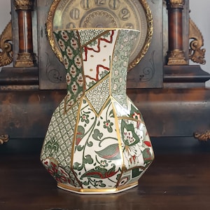 May include: A hexagonal ceramic vase featuring a multi-paneled design with intricate green, white, and red floral and geometric patterns. The panels are separated by thin gold-coloured lines, creating a structured, faceted appearance.