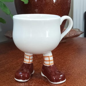 Puede incluir: Taza de cerámica blanca con piernas y zapatos marrones. La taza tiene un patrón de cuadros marrones y naranjas en las piernas.