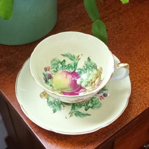 May include: A vintage teacup and saucer set with a delicate floral design featuring a variety of fruits, including apples, grapes, and berries. The cup and saucer are a pale yellow colour with a gold trim.