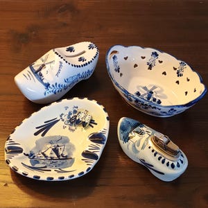 May include: Set of four blue and white Delftware ceramic items, including a shoe-shaped ashtray, a small bowl with a windmill design, a larger bowl with a windmill design, and a shoe-shaped lighter.