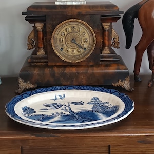 May include: A blue and white ceramic platter with a landscape scene, including trees and birds. The platter has a decorative edge and is placed on a wooden surface. A vintage clock and other ornaments are visible in the background.