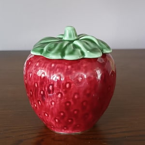 Può includere: Un barattolo in ceramica a forma di fragola con coperchio. Il barattolo è di un rosso vibrante con piccoli urti strutturati e il coperchio è verde, simile a foglie. Il barattolo viene probabilmente utilizzato per riporre piccoli oggetti o come elemento decorativo.
