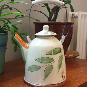 Pode incluir: Uma chaleira de cerâmica branca com uma alça marrom e um design de folhas verdes. A chaleira tem uma tampa branca com uma pequena pega.