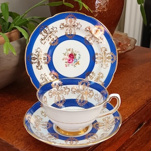 Może przedstawiać: Zestaw do herbaty w stylu vintage, składający się z filiżanki, spodka i talerza. Zestaw ma białą podstawę z niebieskimi i złotymi akcentami. Filiżanka i spodek mają pośrodku kwiatowy wzór. Talerz ma falisty brzeg.