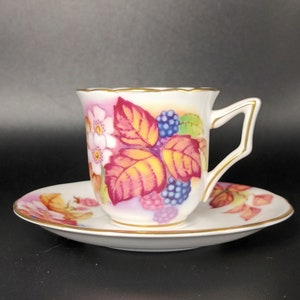 Puede incluir: Una taza de té y un platillo blancos con un diseño floral con flores y bayas rosas, amarillas y moradas. La taza y el platillo tienen un borde dorado.