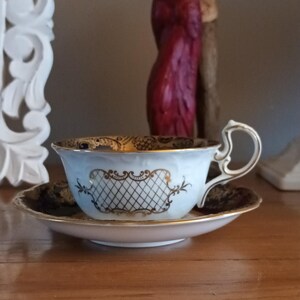 May include: A white teacup and saucer with a blue and gold floral pattern. The cup has a gold trim and a gold lattice design on the front. The saucer has a blue and gold floral pattern on the edge.