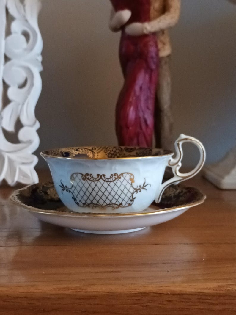 May include: A white teacup and saucer with a blue and gold floral pattern. The cup has a gold trim and a gold lattice design on the front. The saucer has a blue and gold floral pattern on the edge.
