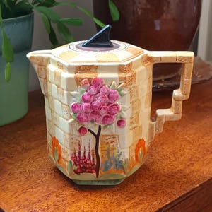 Puede incluir: Una tetera de cerámica vintage con un diseño geométrico y un motivo floral. La tetera está decorada con un patrón de cuadros, un árbol floreciente rosa y flores naranjas y amarillas. La tetera tiene una tapa negra con un pomo puntiagudo.