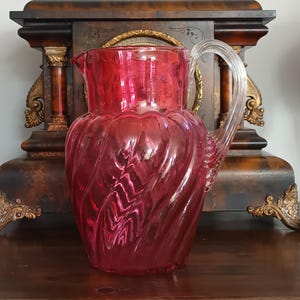 May include: A translucent ruby red glass pitcher featuring a distinct spiral ribbed pattern throughout its body. The vessel includes a clear glass handle with vertical ridges and a wide pouring spout at the top rim, showcasing a vibrant colour.