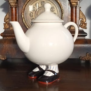 Peut inclure: Une théière en céramique blanche fantaisiste avec un corps rond, un couvercle et un bec verseur. La théière est ornée de chaussures noires et rouges et de chaussettes rayées. La théière est présentée sur une surface en bois devant une horloge vintage.