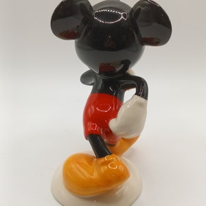 May include: A ceramic figurine of Mickey Mouse standing with his right hand on his hip. He is wearing red shorts, white gloves, and yellow shoes. The figurine is on a white base.