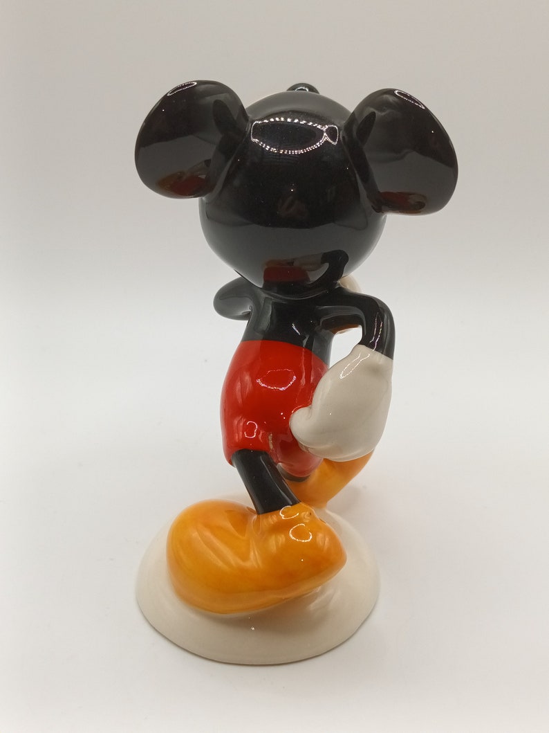 May include: A ceramic figurine of Mickey Mouse standing with his right hand on his hip. He is wearing red shorts, white gloves, and yellow shoes. The figurine is on a white base.