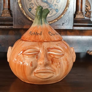 Può includere: Un barattolo di ceramica a forma di cipolla con un volto, con un coperchio con la scritta "Pickled Onion". Il barattolo è principalmente arancione con dettagli verdi e ha un aspetto vintage invecchiato.