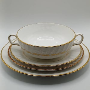 Peut inclure: Un bol à soupe en porcelaine blanche avec une bordure dorée et deux anses, niché dans deux assiettes assorties. Les assiettes ont également une bordure dorée.