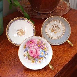 May include: Three vintage teacups with gold trim and floral designs. The cups are white with gold accents and feature different floral patterns. One cup has a blue and gold design, another has a pink and purple floral design, and the third has a gold and white design.