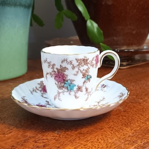 May include: A white teacup and saucer with a floral pattern in shades of pink, brown, and blue. The cup has a gold rim and the saucer has a scalloped edge.