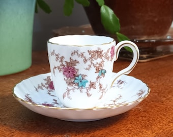May include: A white teacup and saucer with a floral pattern in shades of pink, brown, and blue. The cup has a gold rim and the saucer has a scalloped edge.