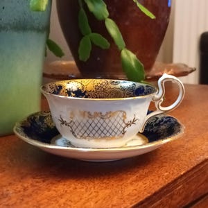 May include: A white teacup and saucer with a blue and gold floral pattern. The saucer has a blue rim and the teacup has a gold handle.