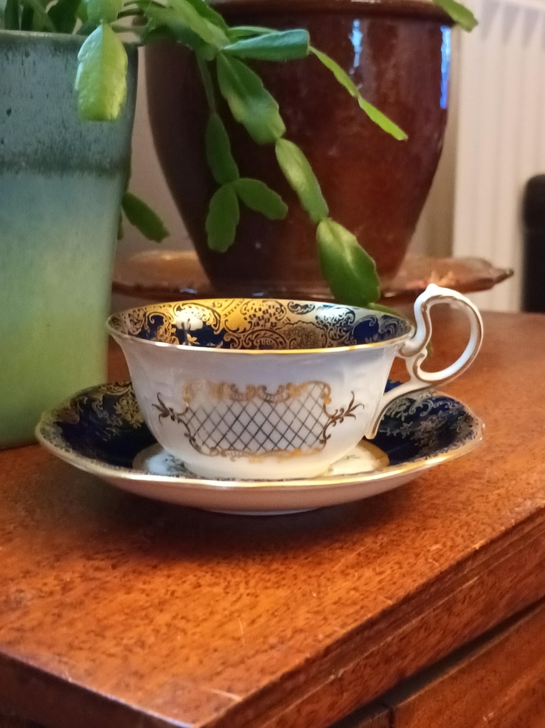 May include: A white teacup and saucer with a blue and gold floral pattern. The saucer has a blue rim and the teacup has a gold handle.