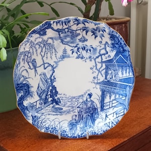 Könnte beinhalten: Ein blauer und weißer Porzellanteller mit goldfarbenem Rand. Der Teller zeigt ein japanisch inspiriertes Design mit einer Pagode, Bäumen und Figuren.