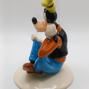 May include: A ceramic figurine of Goofy sitting with his legs crossed, wearing a blue shirt, orange waistcoat, and a yellow hat. He is sitting on a white base.