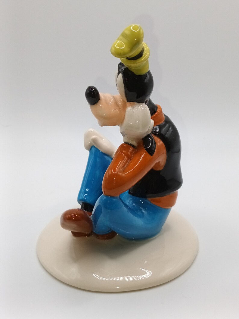 May include: A ceramic figurine of Goofy sitting with his legs crossed, wearing a blue shirt, orange waistcoat, and a yellow hat. He is sitting on a white base.