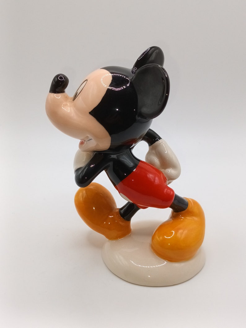 May include: A ceramic figurine of Mickey Mouse in a walking pose. He is wearing his signature red shorts, yellow shoes, and black ears. The figurine is painted in bright colours and has a glossy finish.