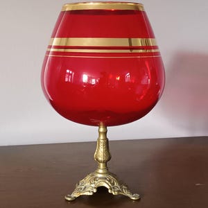 Op de afbeelding: Een rode cognacsnifter met gouden banden en randen, gemonteerd op een decoratieve messing standaard. De bolvormige kom van het glas is helderrood, met twee gouden banden die het midden omsluiten. De standaard heeft een sierlijk, gedetailleerd ontwerp.