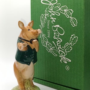 May include: A ceramic figurine of a pig wearing a green vest and holding a pipe. The figurine stands on a green base with grass detail. A green box with the text "John Beswick Since 1894" is in the background.
