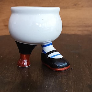 Puede incluir: Un cuenco de cerámica blanco caprichoso con un diseño de pierna y zapato. El zapato es negro con suela roja y detalles a rayas azules y blancas. El cuenco está diseñado para parecer que lleva un zapato.