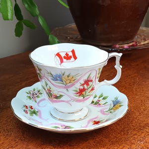 Puede incluir: Taza y platillo de té blancos con un diseño floral y una bandera canadiense en la taza. El platillo tiene los nombres de las provincias canadienses escritos en cintas rosas.