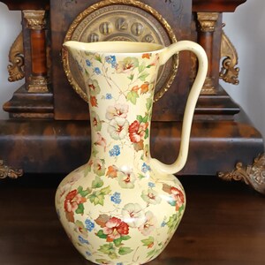 May include: A tall, cream-colored pitcher with a large handle and a floral pattern. The pitcher features a bulbous base and a slender neck. The floral design includes red, pink, blue, and green flowers and leaves on a yellow background.