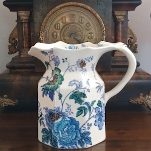 May include: A white ceramic pitcher with a hexagonal body and a scalloped rim, featuring a detailed floral pattern in shades of blue, green, and brown. The pitcher has a curved handle and a pouring spout, decorated with botanical illustrations.