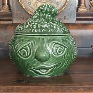 Könnte beinhalten: Ein glänzendes grünes Keramikgefäß mit Deckel in Form einer Figur mit strukturierter Oberfläche. Der Deckel hat ein geformtes Oberteil und der Text "PARSLEY SAUCE" ist vorne eingeprägt. Das Gefäß hat einen runden Boden mit einem Gesicht.