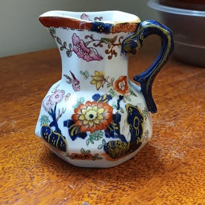 May include: A small, white ceramic jug with a blue and gold floral pattern. The jug has a blue handle with gold accents.