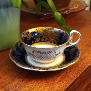 Könnte beinhalten: Eine weiße Teetasse und Untertasse mit einem blauen und goldenen Blumenmuster. Die Tasse hat einen goldenen Rand und ein zartes Design an der Seite. Die Untertasse hat ein passendes Muster.