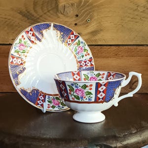 Puede incluir: Juego de taza y platillo de té blanco con un patrón floral y geométrico en rojo, azul y dorado. La taza tiene un asa y se apoya en una pequeña base. El platillo es redondo y tiene un diseño a juego.