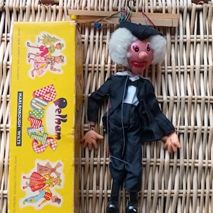 Puede incluir: Una marioneta Pelham vintage con un traje negro, camisa blanca y pajarita, con peluca blanca y cara roja. Junto a la marioneta, una caja amarilla con ilustraciones y el texto "Pelham Marlborough Wilts".