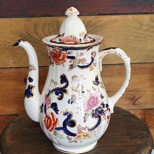 Puede incluir: Una tetera de cerámica blanca con tapa, con diseños florales en rojo, rosa y azul. La tetera tiene un pico y asa curvos, con ribetes dorados. Está colocada sobre un pequeño taburete de madera.